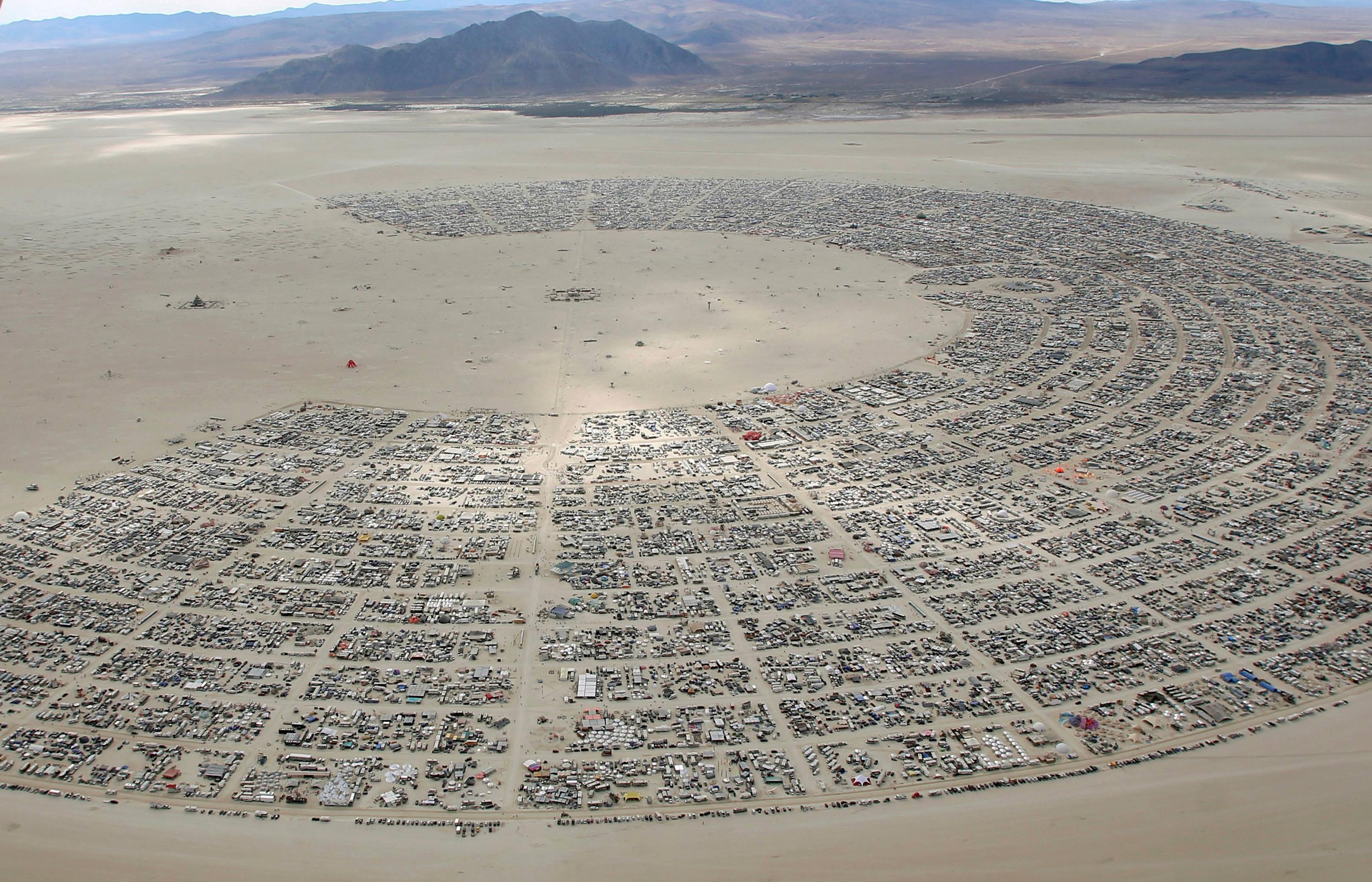 The meme is an image of the Burning Man festival in the Black Rock Desert, with a large crowd of people and a sea of cars and RVs. The text "I love you" is written in the sand, but it's being erased by a giant foot, implying that the love is being destroyed by the festival's chaos and excess.